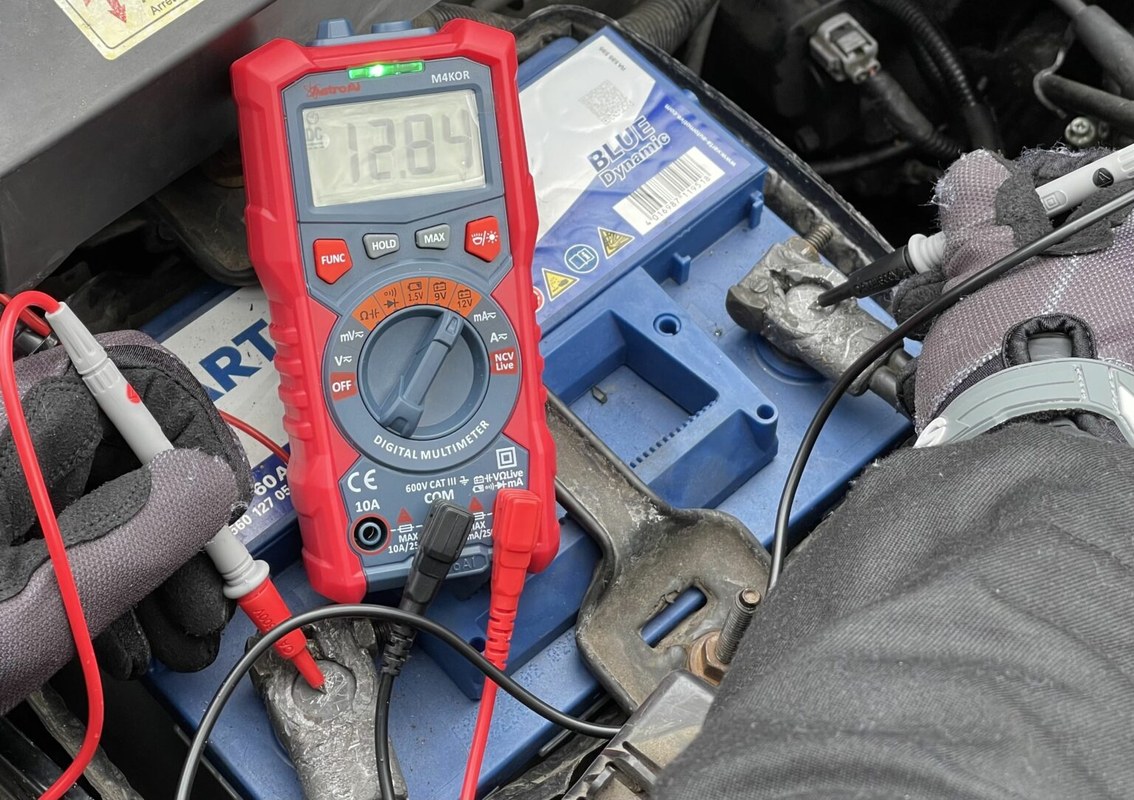 Person testing a car battery safely with a multimeter and battery tester under an open hood