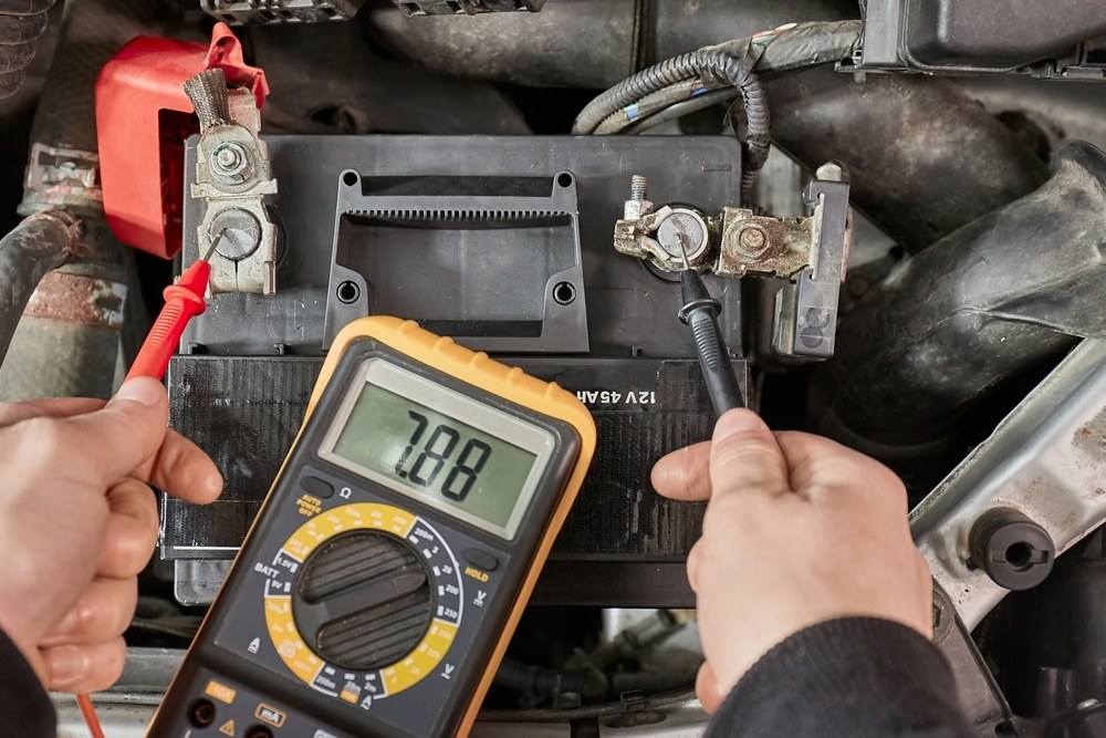 Multimeter probes connected to a car battery terminal with safety gloves and goggles nearby